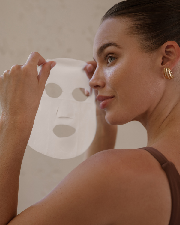 Woman holding a sheet mask to her face against a neutral background